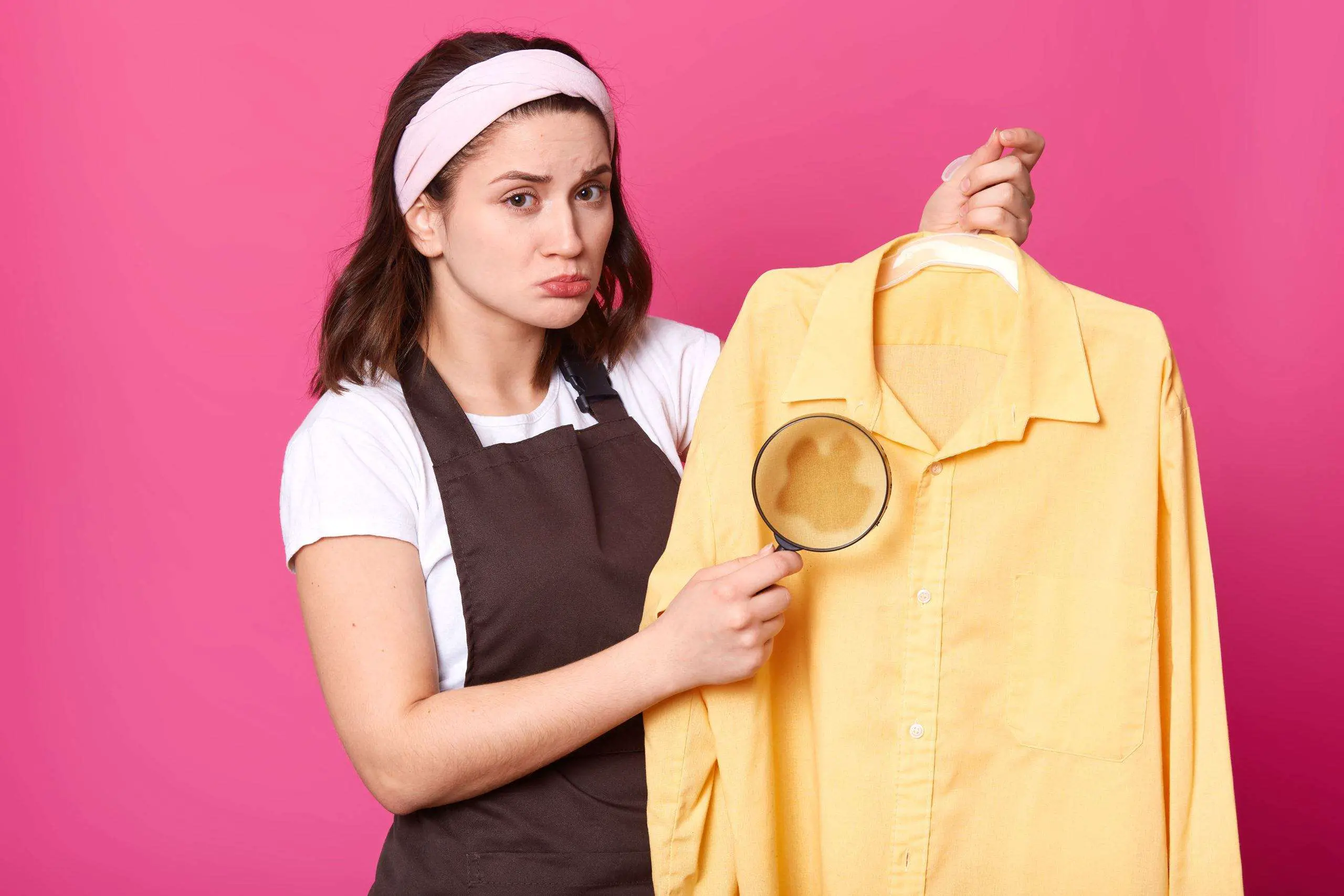 Brunette housewife holds yellow shirt after laundry with coffee stain on fabric, having magnifying glass in one hand. Emotional cute young model wearing brown apron, white t shirt and headband.